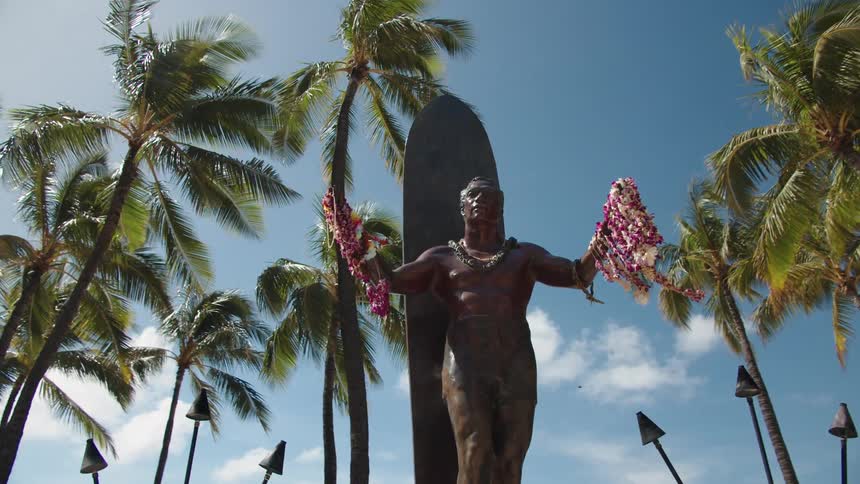 Bronze sculpture of man against a surfboard holding leis at a beach resort