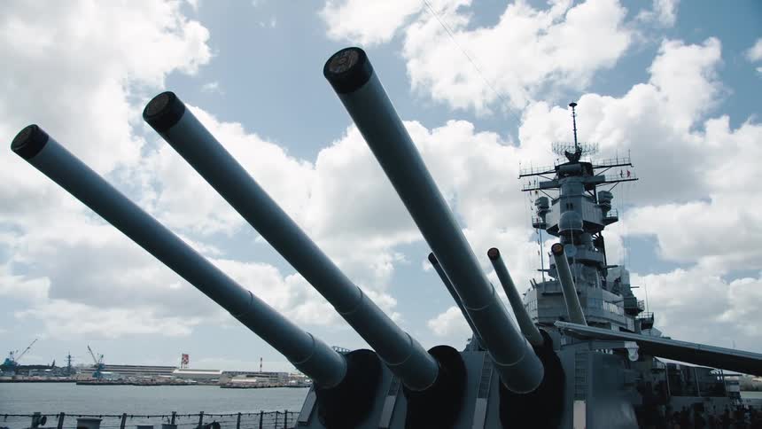 People visit navy vessel with guns docked across from warehouses and cranes