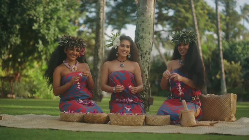 Three young pacific islander women string leis on blanket