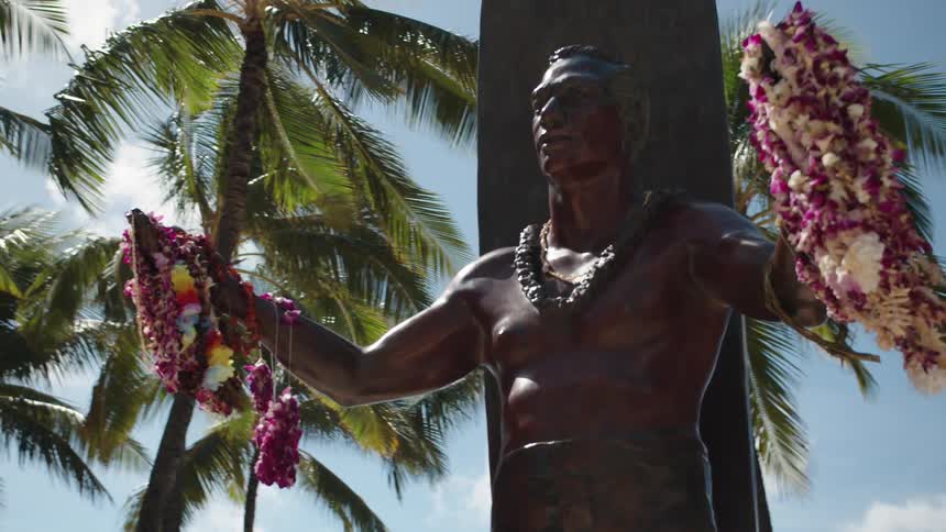 A statue of a man holding leis with trees and the sky in the background