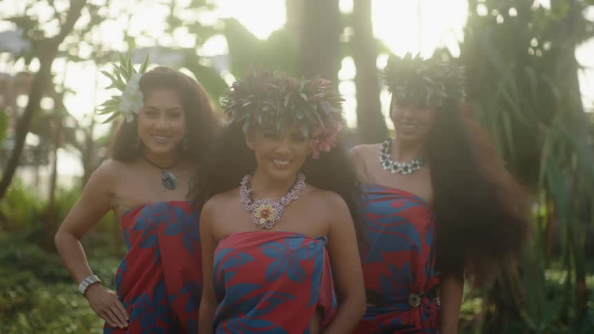 Three hawaiian women wearing the same dress and flowers in their hair pose and smile while the wind blows their hair
