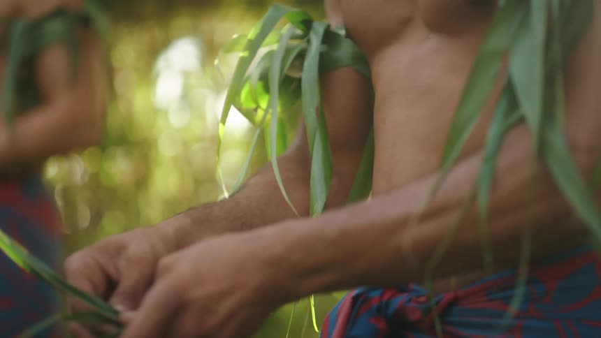 Two men wearing woven headbands weave plant material together