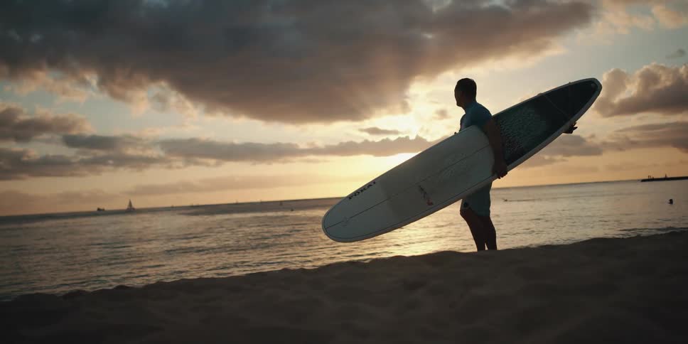 A man standing on the beach then bending down and picking up a surfboard
