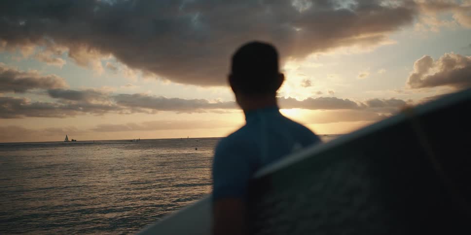 A surfer holding a surfboard looking out over the ocean towards the low sun