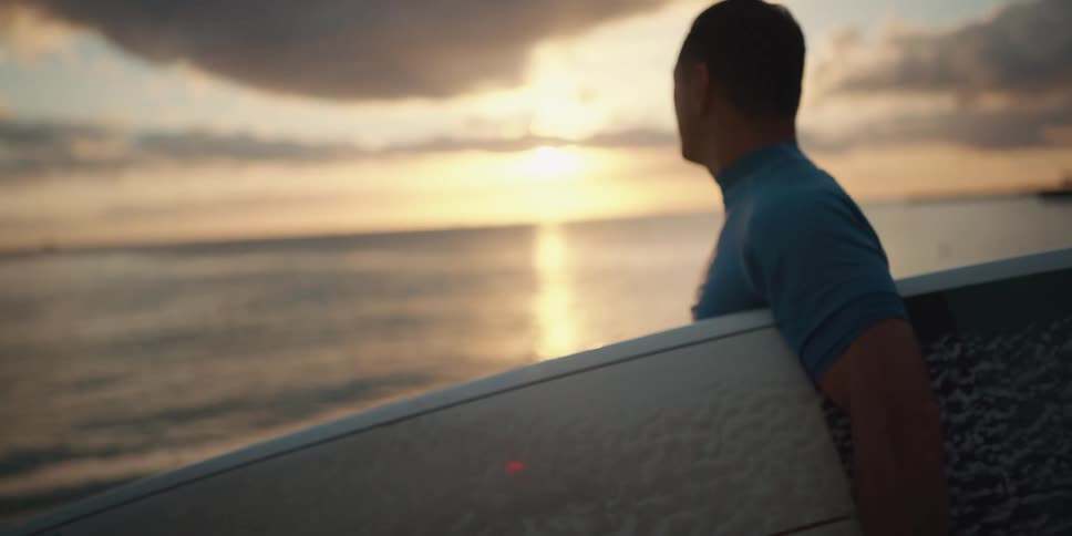 Male surfer picking up his surfboard then looking out at ocean at sunset