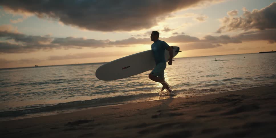 Man with surfboard runs along beach at sunset