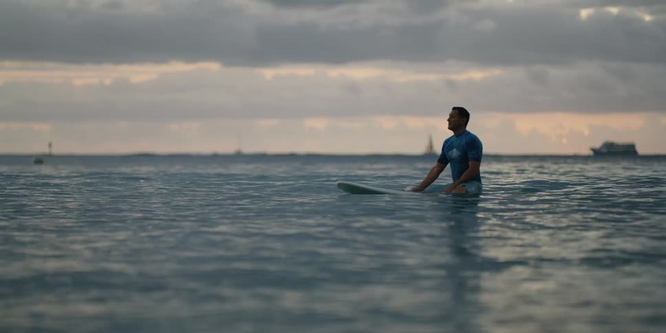 A man sits on his surfboard floating in the ocean at sunset