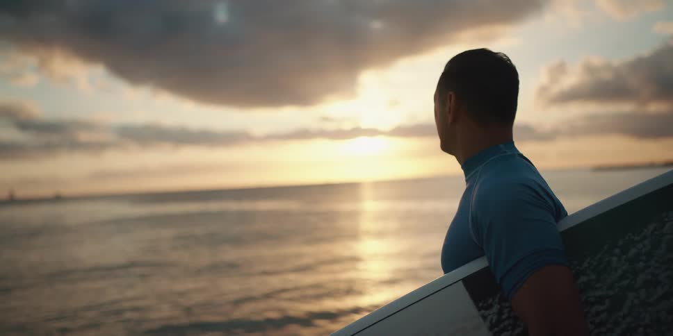 Man standing on the white sand beach picks up the surfboard and watches sunset
