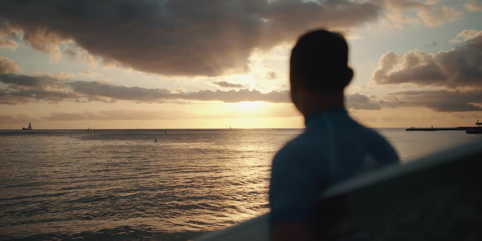 A man holding a surfboard looks out over the ocean at sunset