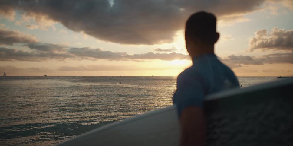 A man holding a surfboard stands on a beach by the ocean at sunset