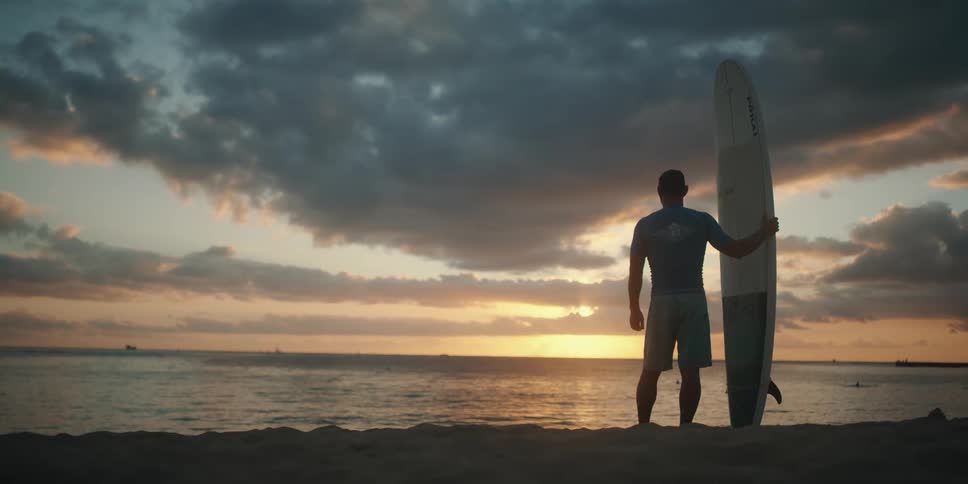 A man stands on the shore holding his surfboard and looking out at the water