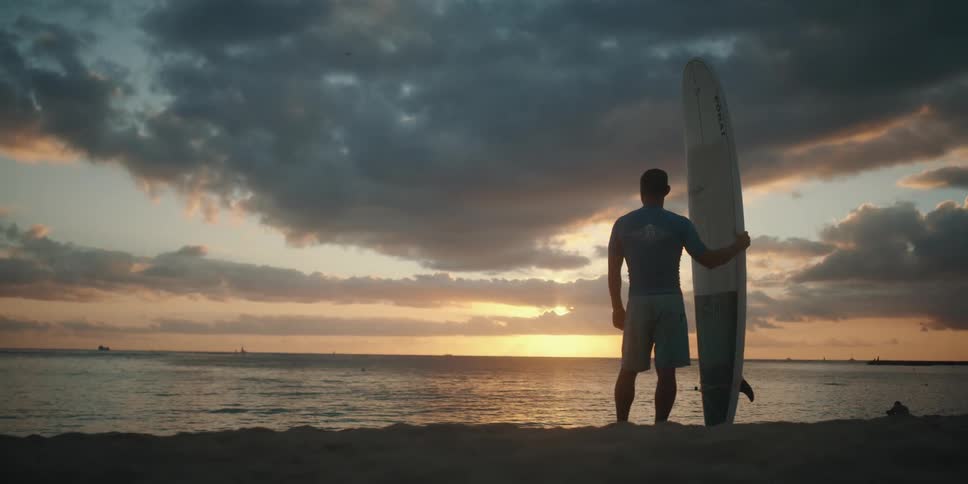 A man holding a surfboard stands on a beach at sunset