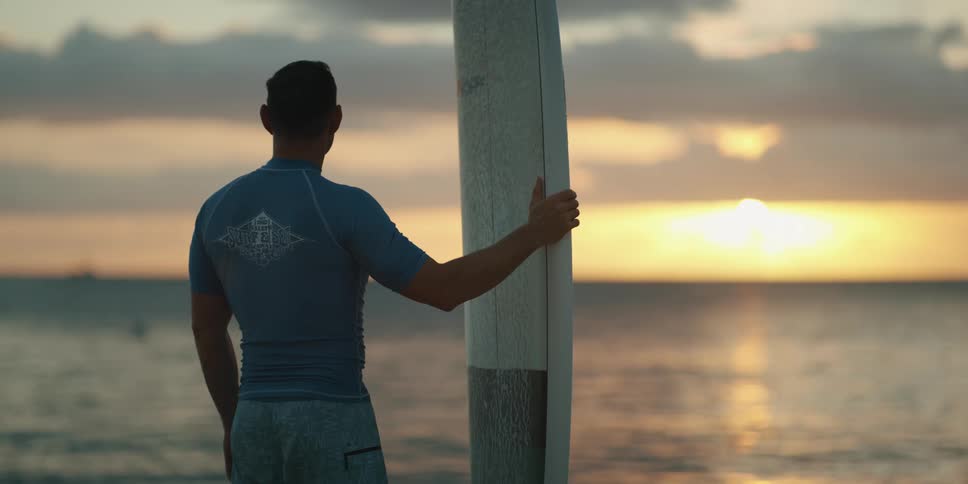Man with surfboard stands on beach looking at sunset