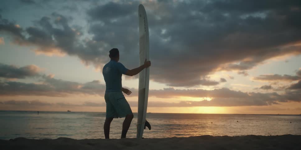 Man holding surfboard looks at sunset from beach