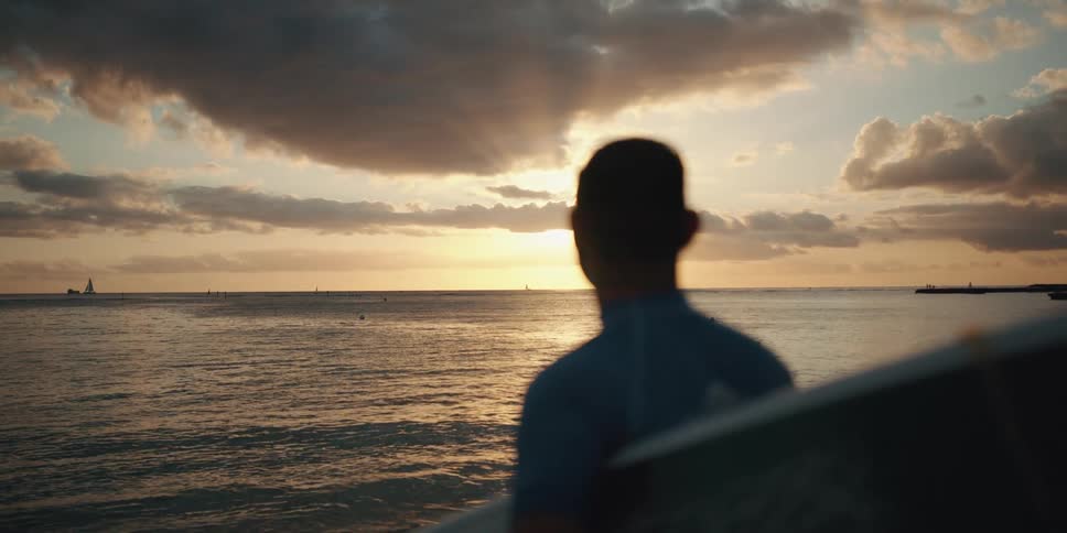 Man with surfboard looks across ocean towards sunset