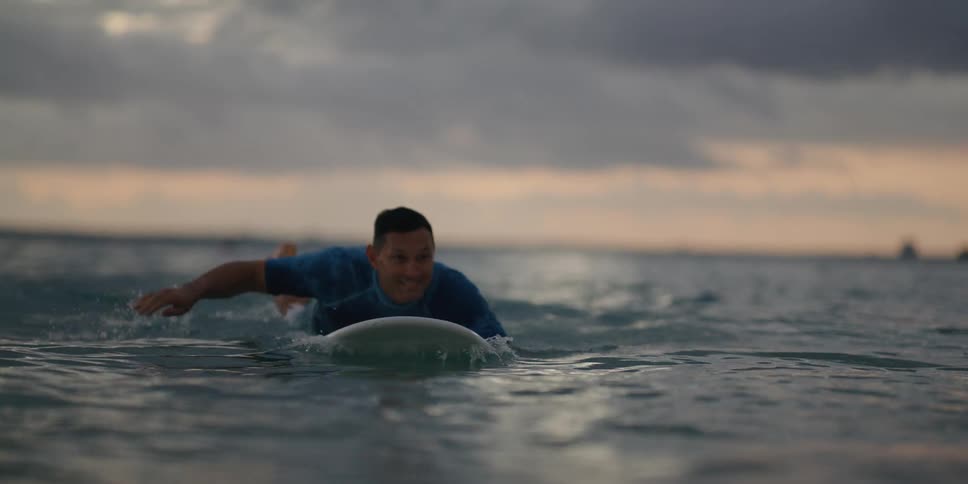 A man paddles a surfboard to the shore from the ocean at sunset