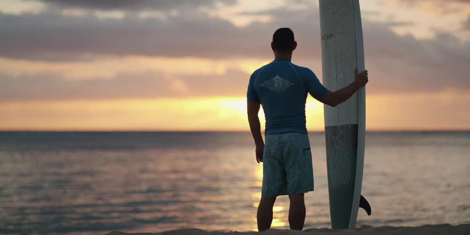 Man standing on the sand beach holds a surfboard and watches the sunset