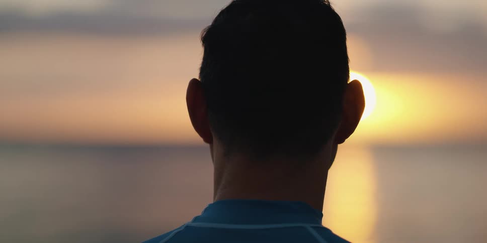 Man with surfboard stands on the beach looking at sunset