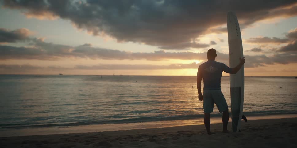 Man stands on the beach holding surfboard looking at sunset over the sea