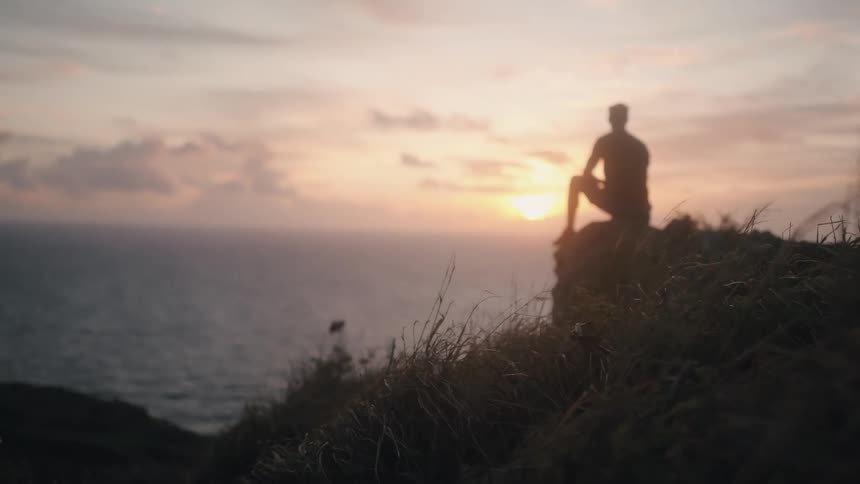 Man sits on rock overlooking ocean at sunset