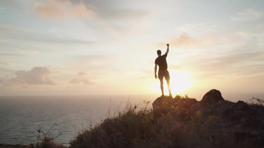A man standing on a cliff near the coast raising his fist to the sky