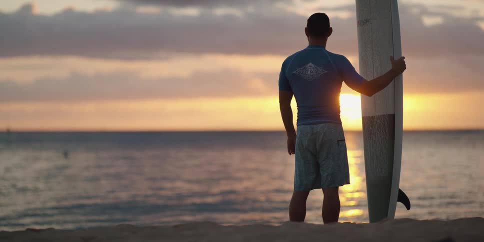 Man standing with surfboard looking out to the ocean at sunset