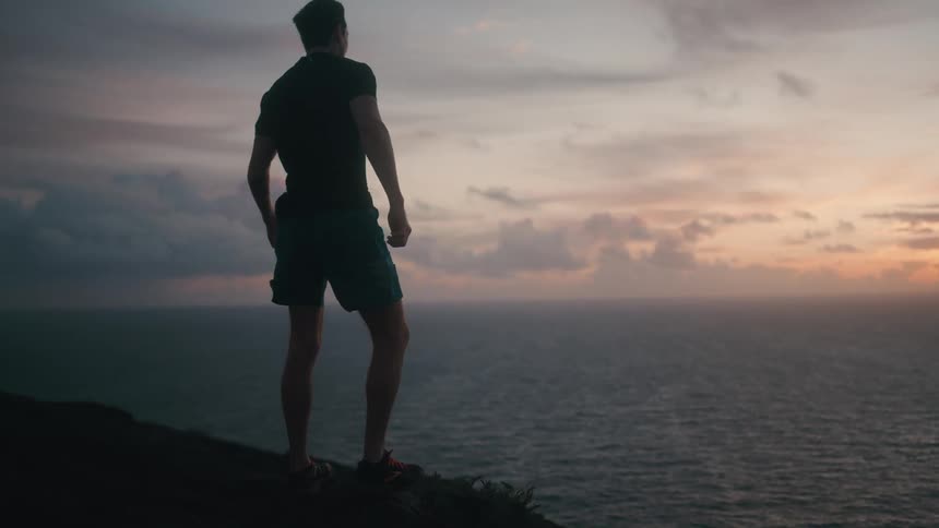 Man jogs up incline to stand and look at ocean view as the sky near the horizon is tinged red