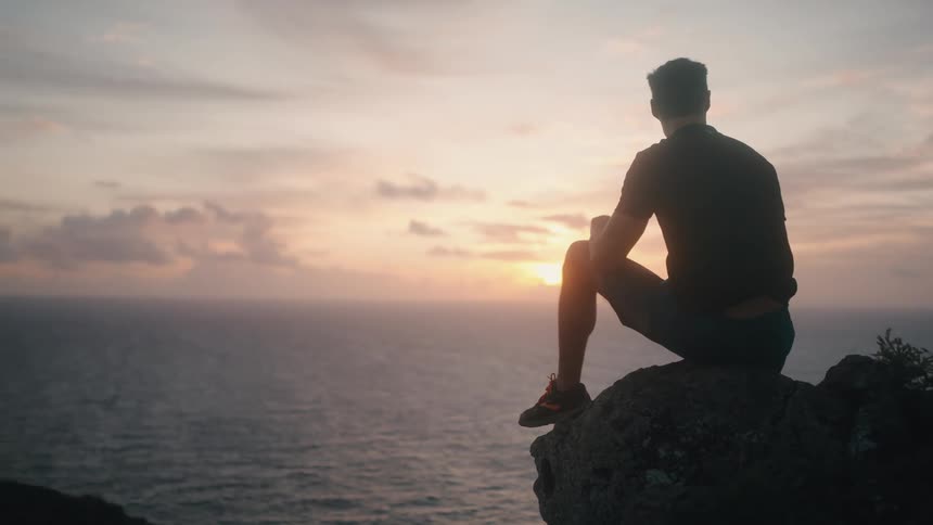A man is sitting on a cliff watching the sunset above the ocean