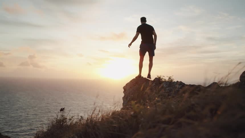 Man jogging to end of cliff at sunrise and celebrating while looking out ocean