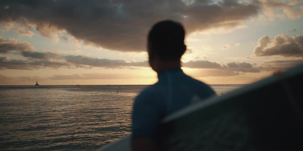 Man with surfboard looks across ocean towards sunset