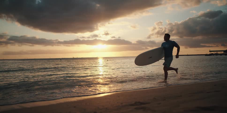 Sun hovers near the horizon as man runs across beach carrying surfboard