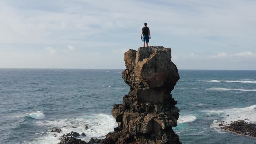 Man climbs rock on beach and stands on it looking at the ocean