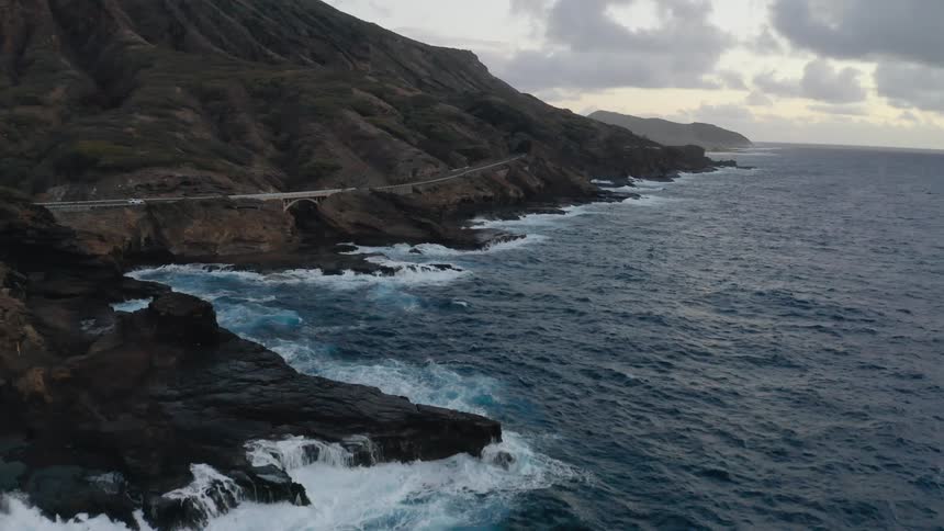 Aerial of waves crashing on shoreline near a road on the mountainside