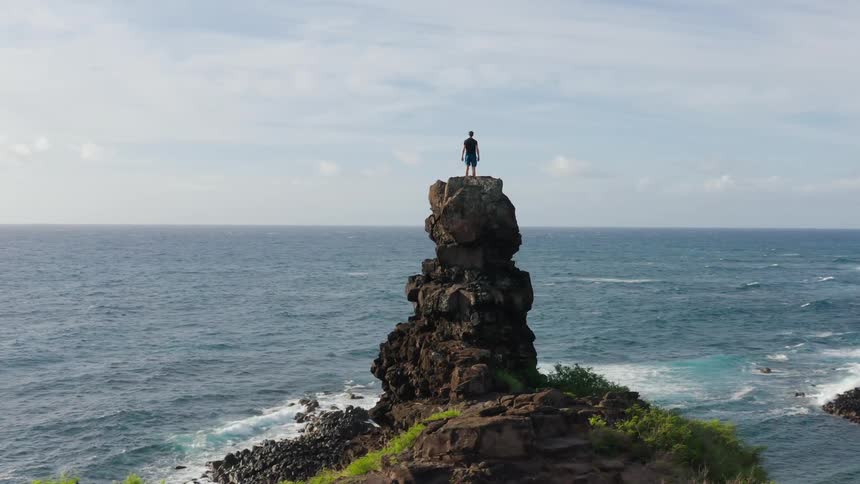 Aerial of a man climbing large rock then standing and looking out to ocean