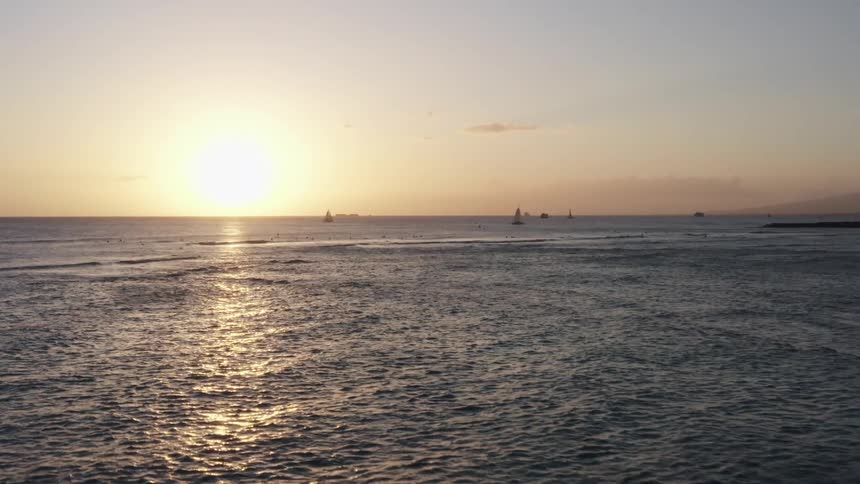 Surfers and swimmers in the waves with sailboats behind at sunset