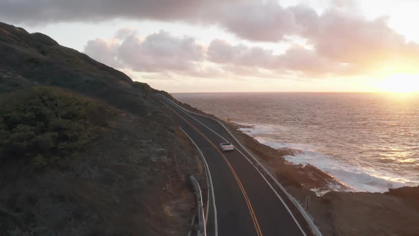 A car travels down a two lane road winding along the coast at dusk