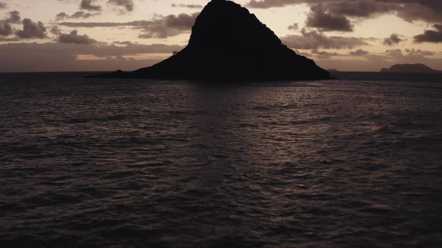 Ocean waves and surface reflect the last of the sunlight as a rocky island comes into view