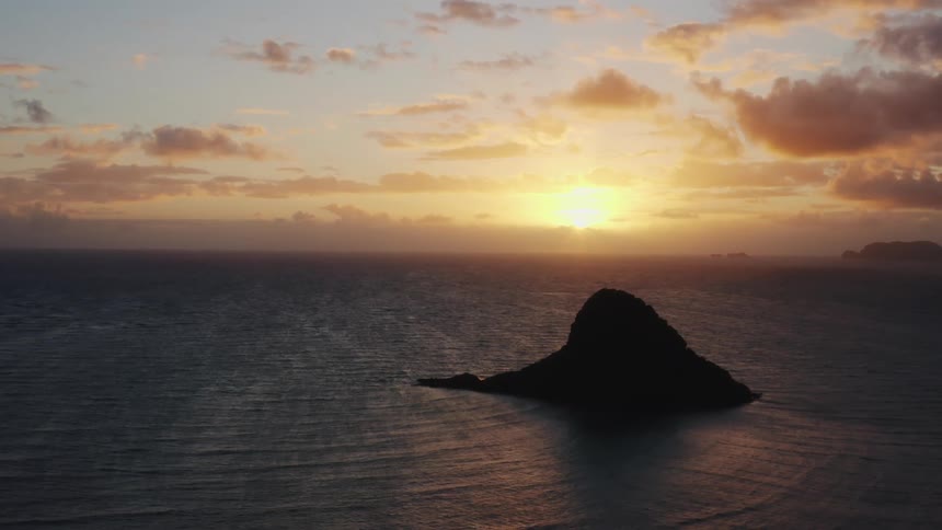 Sun rises over waves forming concentric rings as they pass near rocky islet