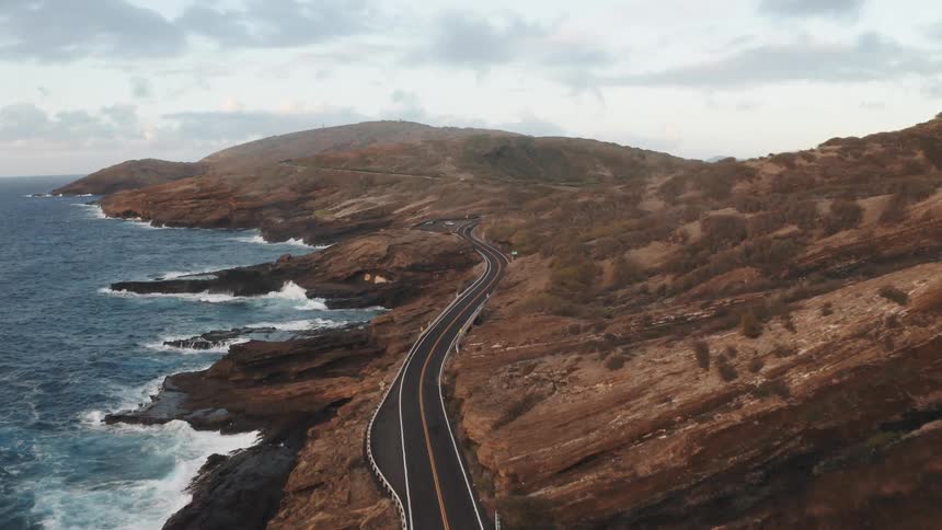 Aerial view of ocean waves crashing against big rocks and mountain highway