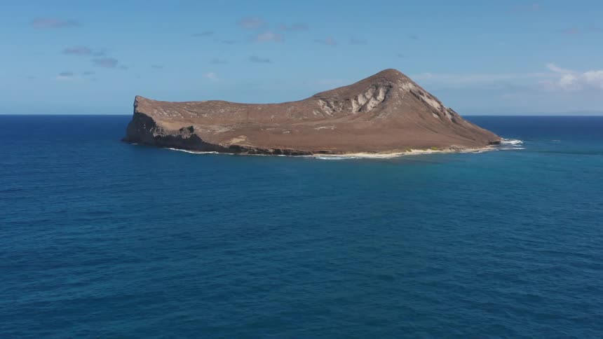 Vegetation free grey island surrounded by blue ocean under a lightly cloudy sky