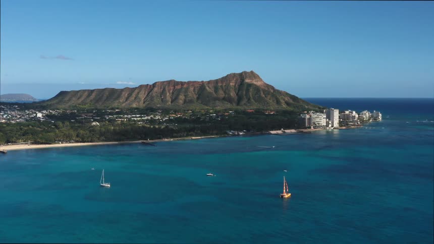 Sailboats sail near city occupying shore next to mountain
