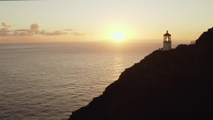 A lighthouse overlooks an ocean with the sun setting on the horizon