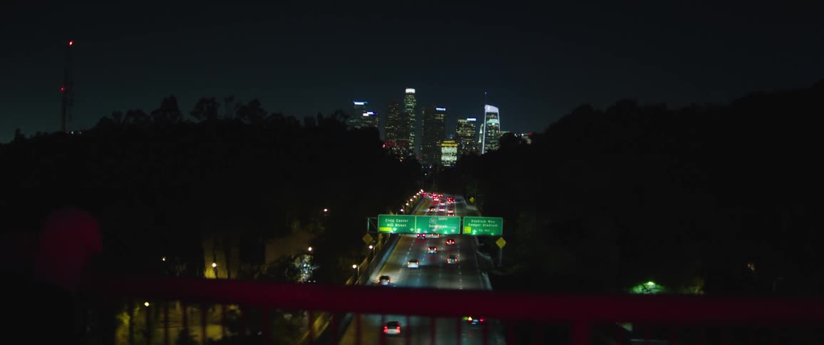 Couple leans against rail overlooking traffic traveling toward downtown skyline at night