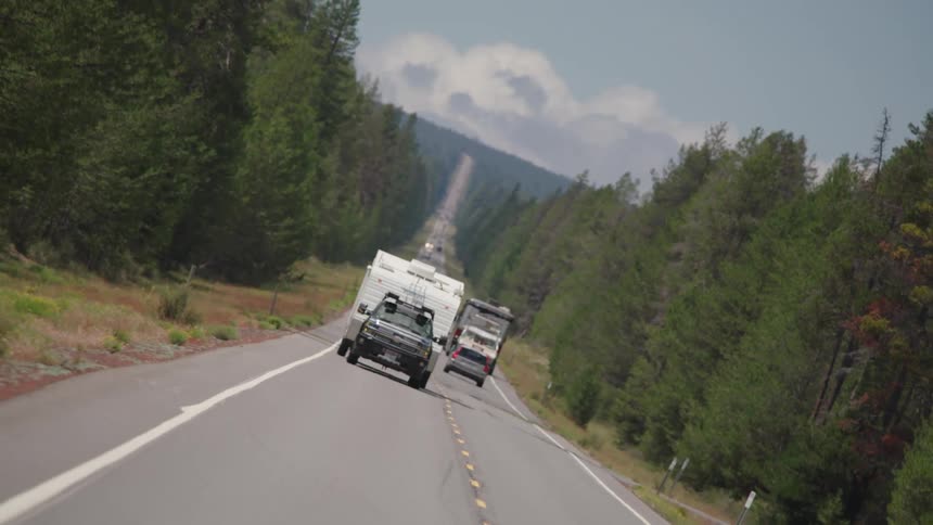 A truck pulling a camper drives down a road through a forest as other cars drive in the opposite direction