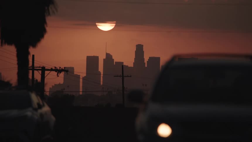 Sun floats in red sky above skyline as cars drive away