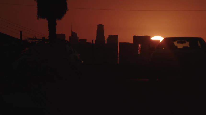 Sun sets behind city skyline with silhouetted cars driving ahead