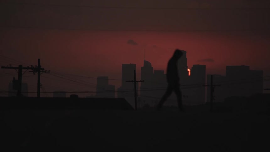 A man wearing a hoodie walks across the street as the sunsets behind the city skyscraper in a glowing pink red sky