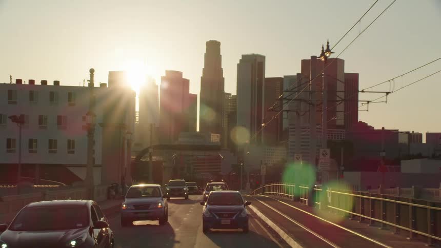 The sun shines through skyscrapers while the vehicles drive along the highway in the city in the late afternoon