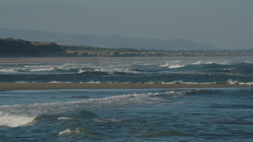 Wave roll in crashing on sand bar from both sides as fog shrouds mountains behind shore