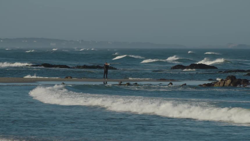 Man standing on the wet sand beach takes picture of the ocean waves next to the sea